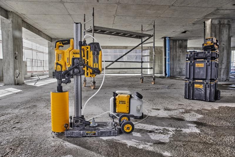 Maszyna do wiercenia rdzeniowego DEWALT z żółtym cylindrycznym wiertłem i podłączonym zbiornikiem na wodę znajduje się na podłodze betonowego placu budowy. W tle widoczny jest rusztowanie oraz układane w stos skrzynie DEWALT.