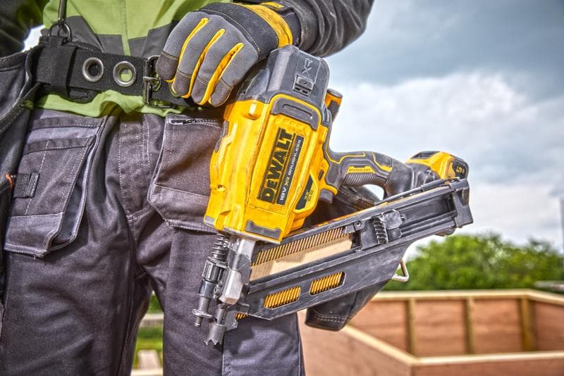 Person holding a DEWALT DCN930N nail gun, wearing work gloves and gear at a construction site outdoors.
