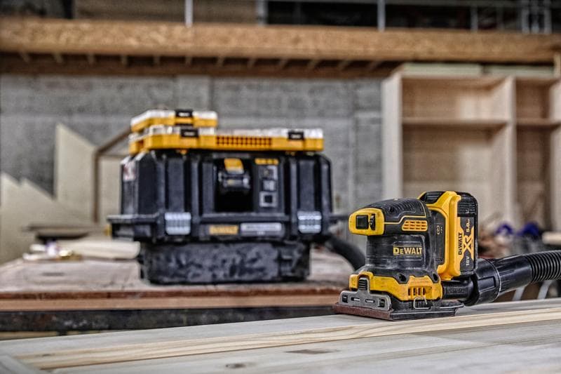 DEWALT cordless sander on wood, with storage box in background in a workshop setting.