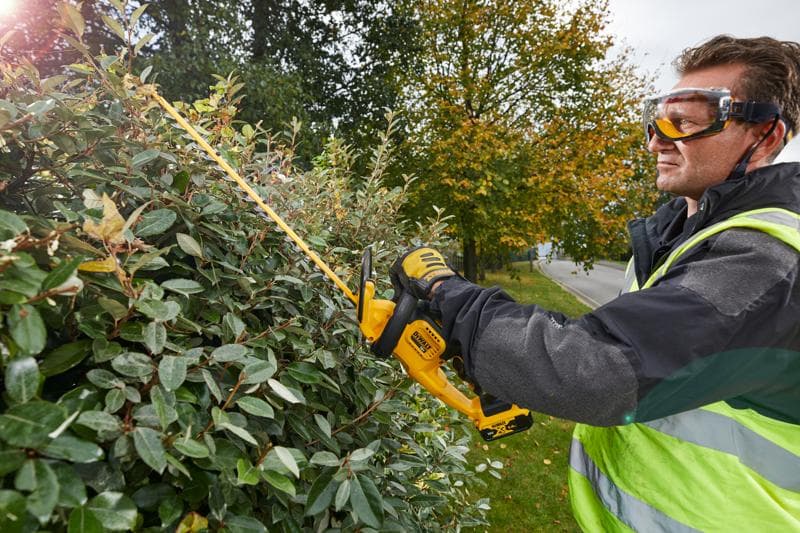 Osoba w sprzęcie ochronnym i rękawicach używa bezprzewodowego nożyc do żywopłotu DEWALT DCM563PB do przycinania gęstego krzewu na zewnątrz. Twarz osoby jest zamazana dla zachowania prywatności.
