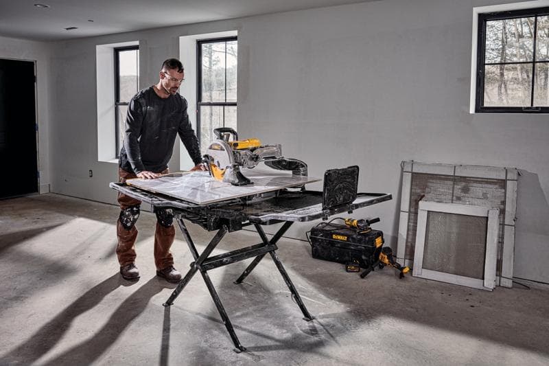 A person stands beside a DEWALT D36000S tile saw setup on its stand in a room under construction. The saw is placed on a foldable black stand, and there are tools and a DEWALT tool bag on the floor nearby. The room has unfinished walls, windows, and construction materials leaning against the wall.