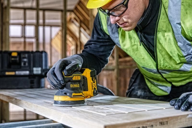 DEWALT DCW210, DCW210D1 sander in use on wood in construction scene. Person is wearing gloves and safety vest.