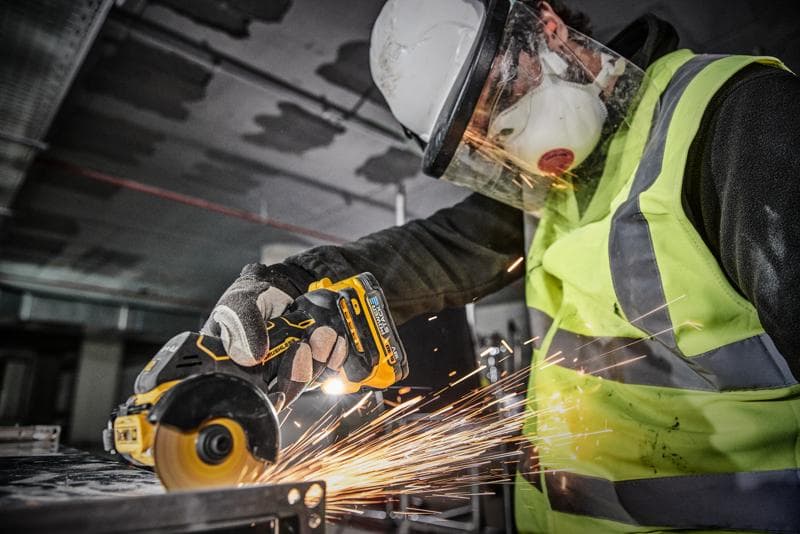 Osoba w sprzęcie ochronnym, w tym kasku, przyłbicy, masce, rękawicach i kamizelce odblaskowej, używa bezprzewodowego narzędzia DEWALT DCS438N do cięcia metalu, co powoduje powstawanie iskier.