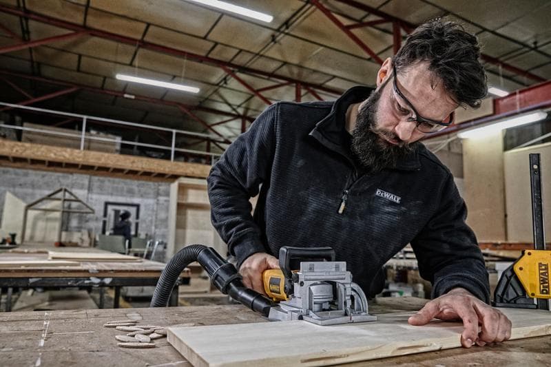 A person using a DEWALT DW682K woodworking tool to cut a piece of wood on a table in a workshop. The person's face is obscured for privacy.