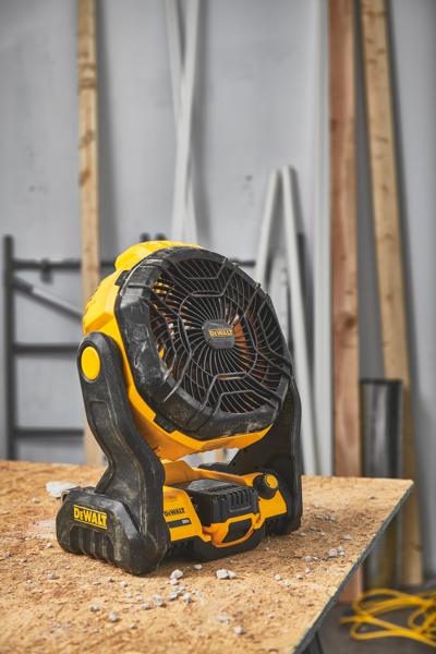 Przenośny wentylator akumulatorowy DEWALT (model DCE512B) stoi na drewnianym stole warsztatowym w warsztacie z materiałami budowlanymi w tle.